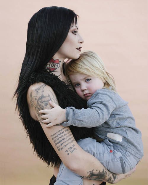 a woman holding a small child with tattoos on her arms and chest, while she is wearing an angel wings jacket