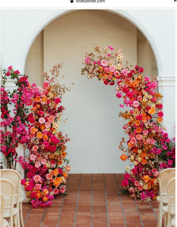 an archway decorated with pink and orange flowers in front of a white wall filled with chairs