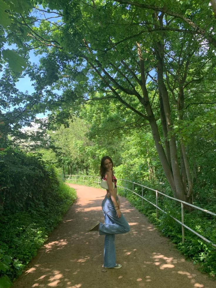 a woman standing in the middle of a dirt road with trees on both sides and a fence behind her