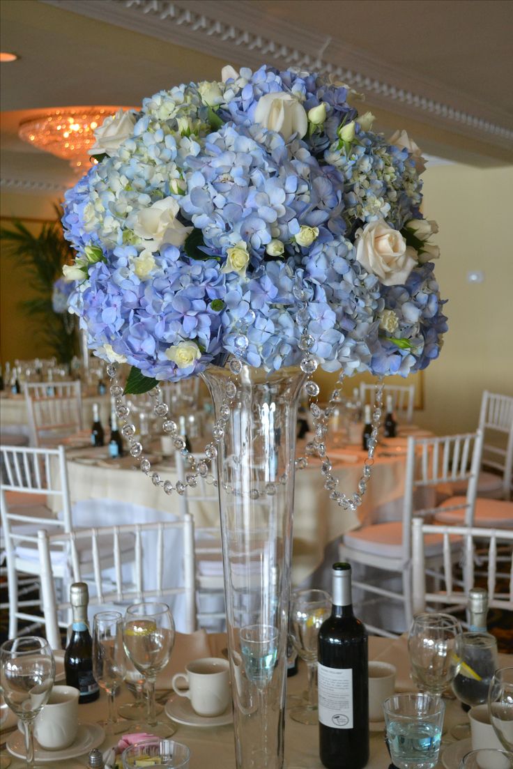 a tall vase filled with blue and white flowers on top of a table next to wine glasses