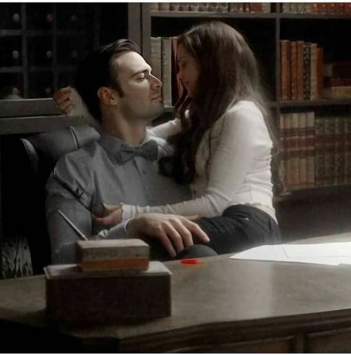 a man and woman sitting at a desk in front of a book shelf with books