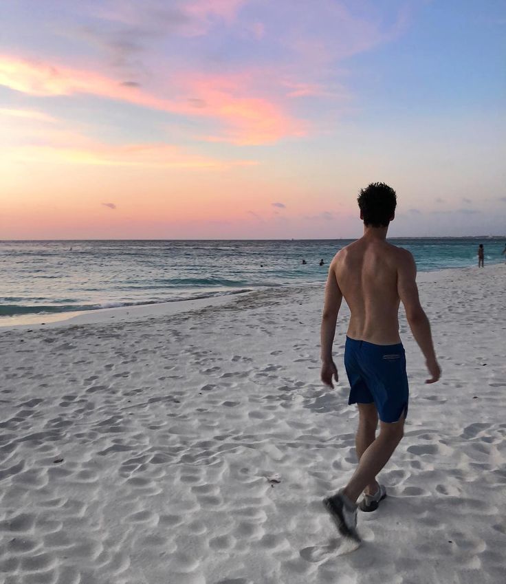 a shirtless man walking on the beach with his feet in the sand as the sun sets