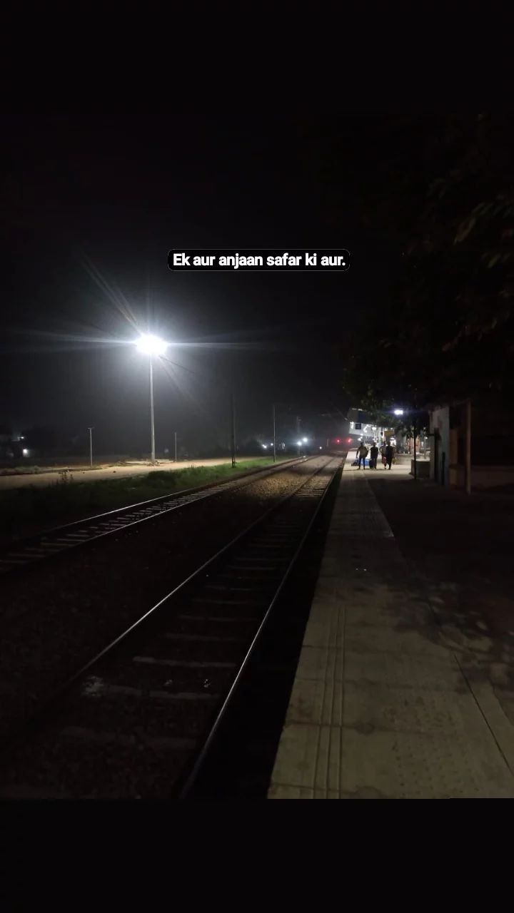 an empty train track at night with people walking on the platform and lights in the background