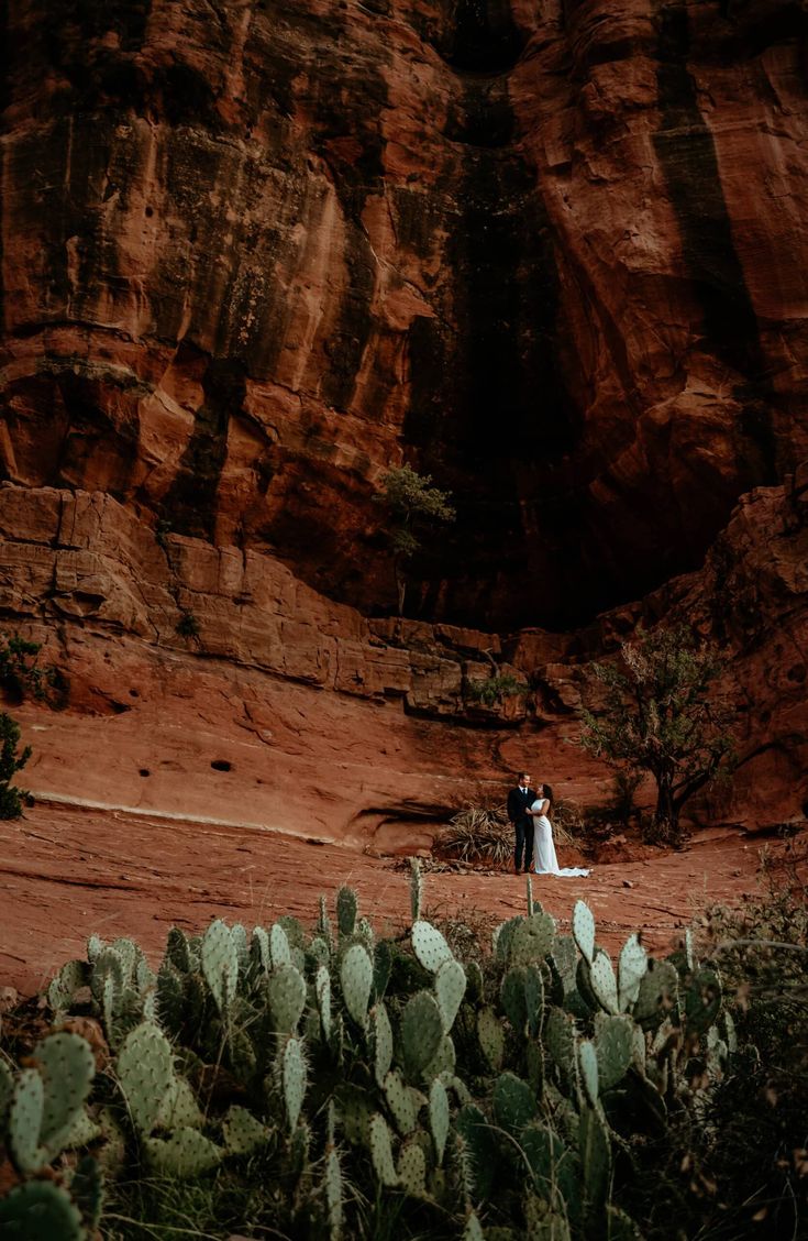 a bride and groom standing in front of a rock formation at the base of a cliff