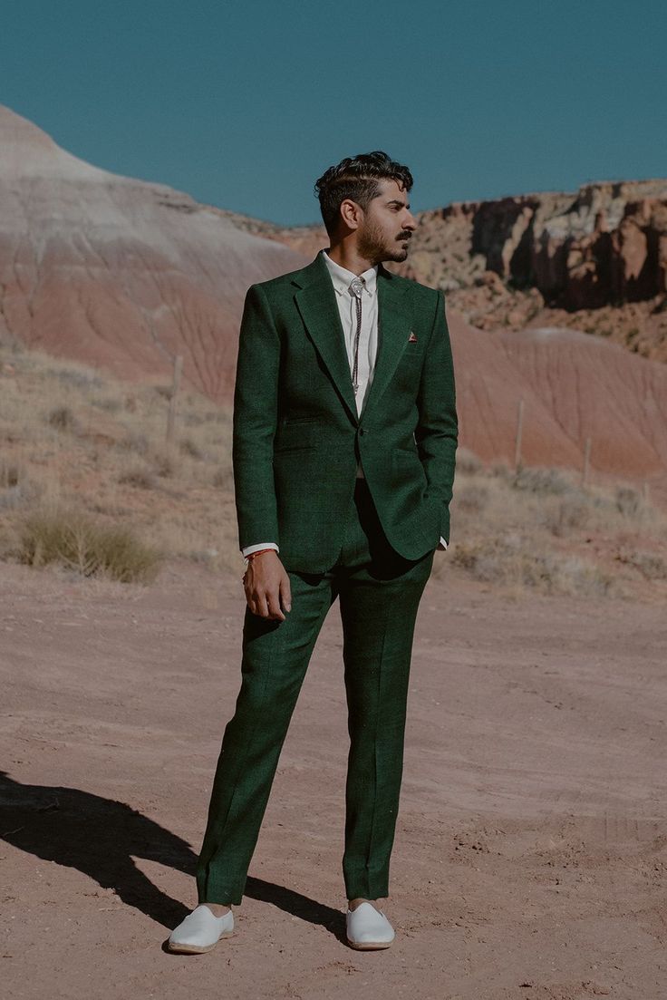 a man in a green suit and white shirt is standing on a dirt field with mountains in the background