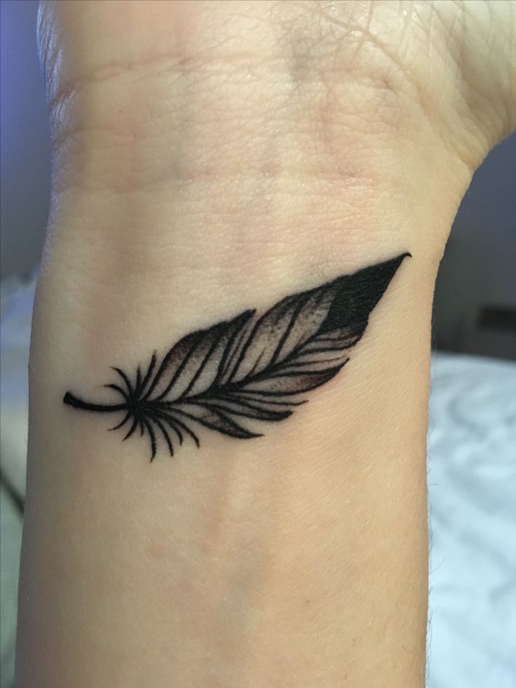 a small black feather tattoo on the wrist is shown in close up, it appears to be an arrow