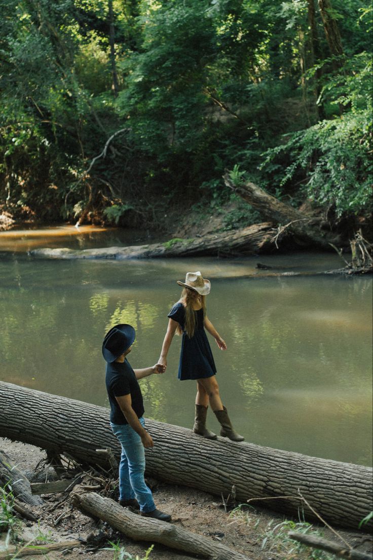 two children holding hands while standing on a log in front of a river and trees