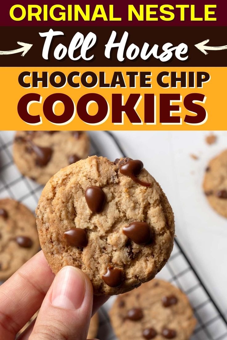 a hand holding up a chocolate chip cookie in front of other cookies on a cooling rack