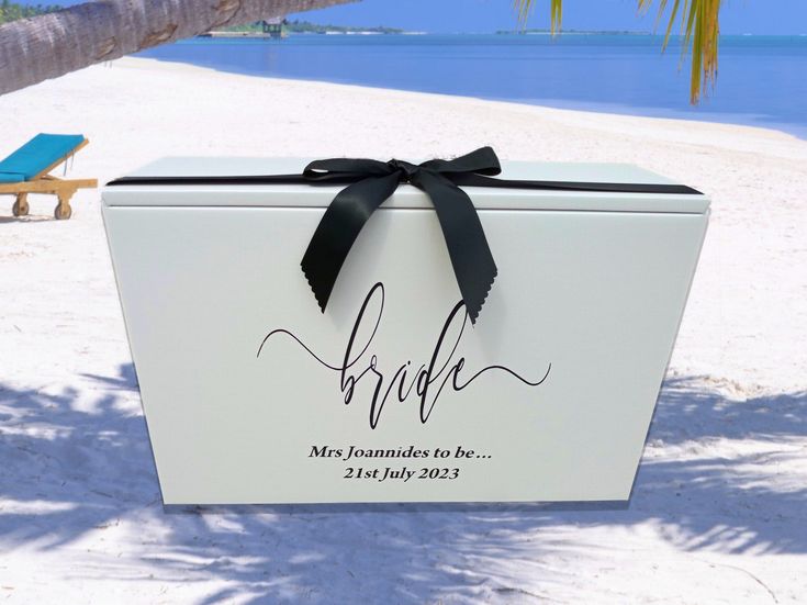 a white box with a black ribbon on the beach next to a chair and palm tree