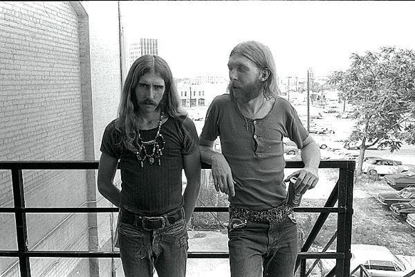 two men standing next to each other on a balcony near a brick building and trees