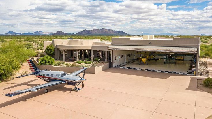 an airplane is parked in front of a building with mountains in the background and clouds in the sky