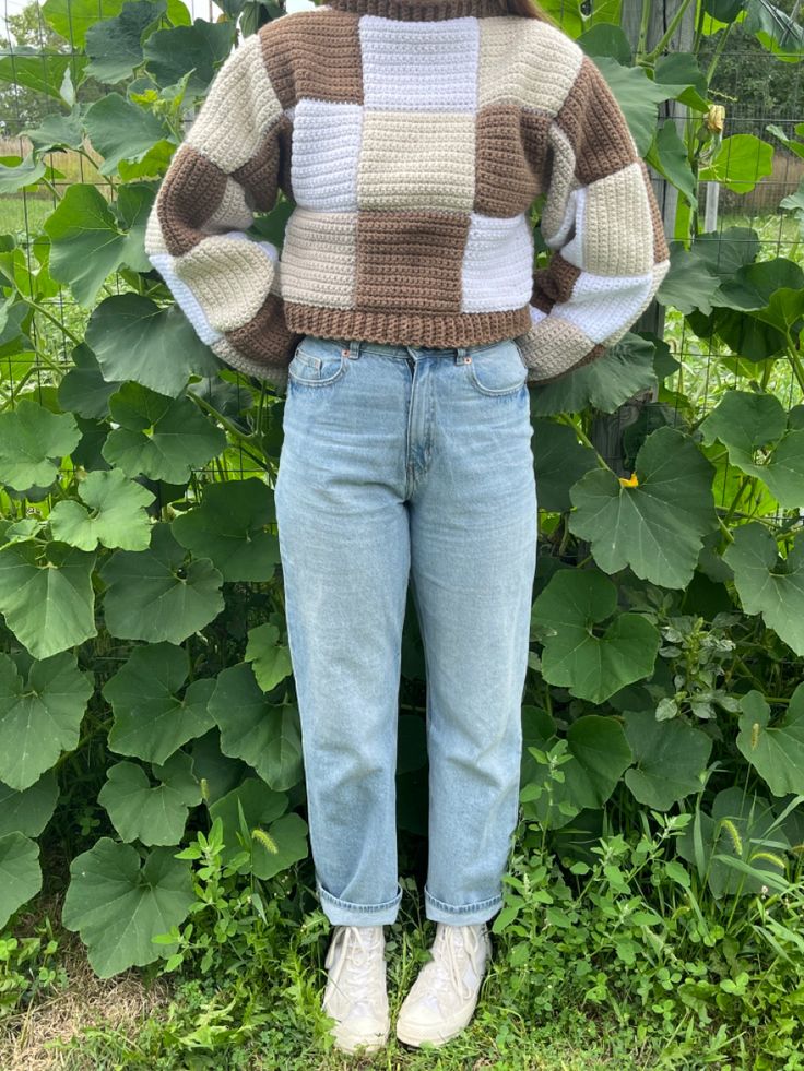 a woman standing in front of green plants wearing jeans and a sweater with squares on it