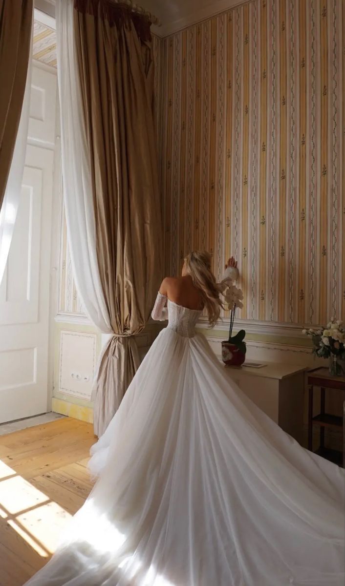 a woman in a white wedding dress is standing near a window with curtains on it