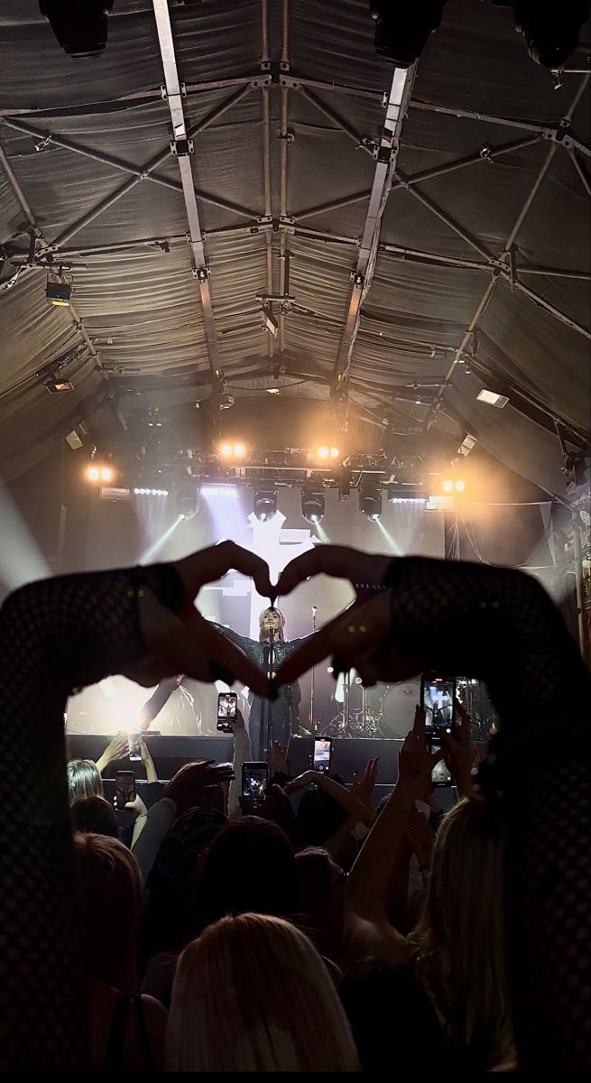 two hands making a heart shape in front of an audience at a music concert with lights on the ceiling