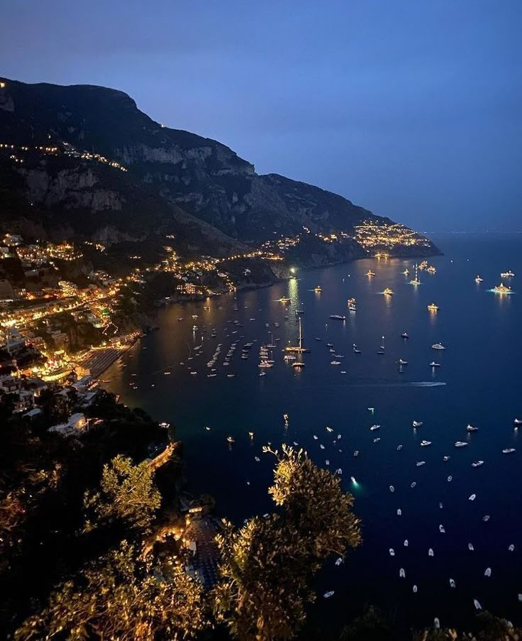 boats are floating in the water at night near some mountains and lights on the shore