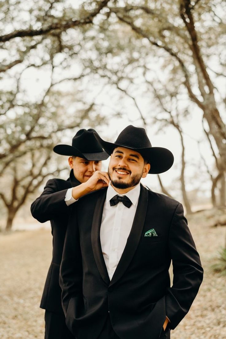 two men in black suits and hats are posing for the camera with trees in the background