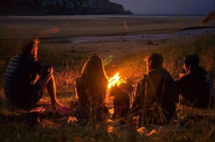 four people sitting around a campfire at night on the beach with an island in the background