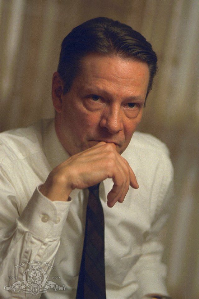 a man in a white shirt and tie sitting at a table with his hand on his chin