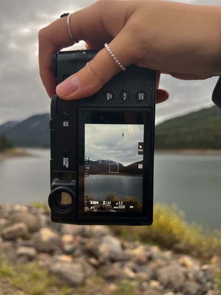 a person is holding up a camera to take a photo near a lake with mountains in the background