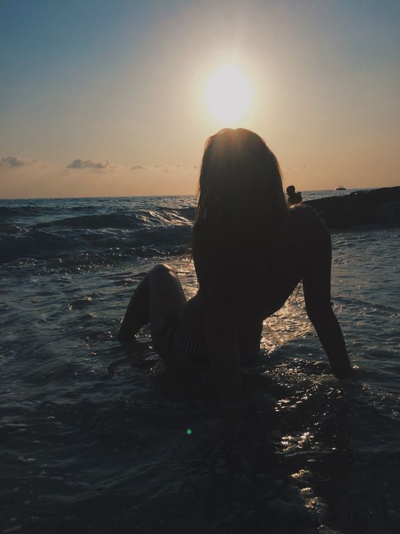 a woman sitting in the ocean at sunset with her back to the camera and arms behind her head