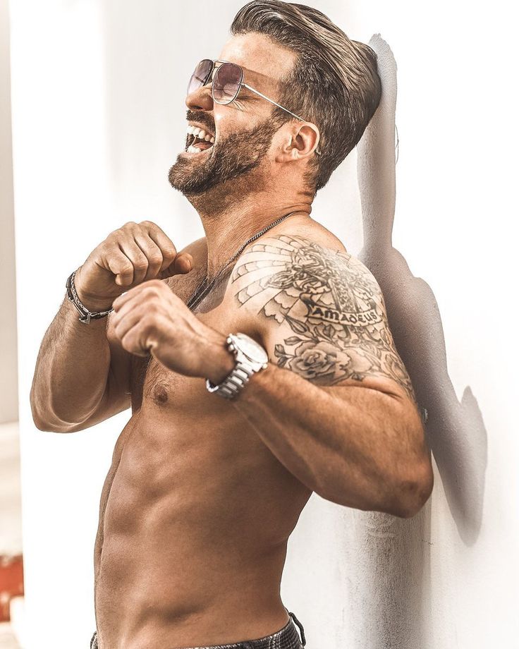 a shirtless man with his arm around his neck and sunglasses on, leaning against a wall