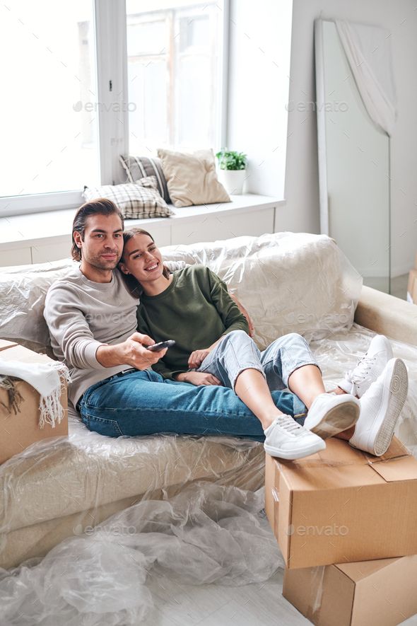 a man and woman sitting on a couch in their living room with boxes around them