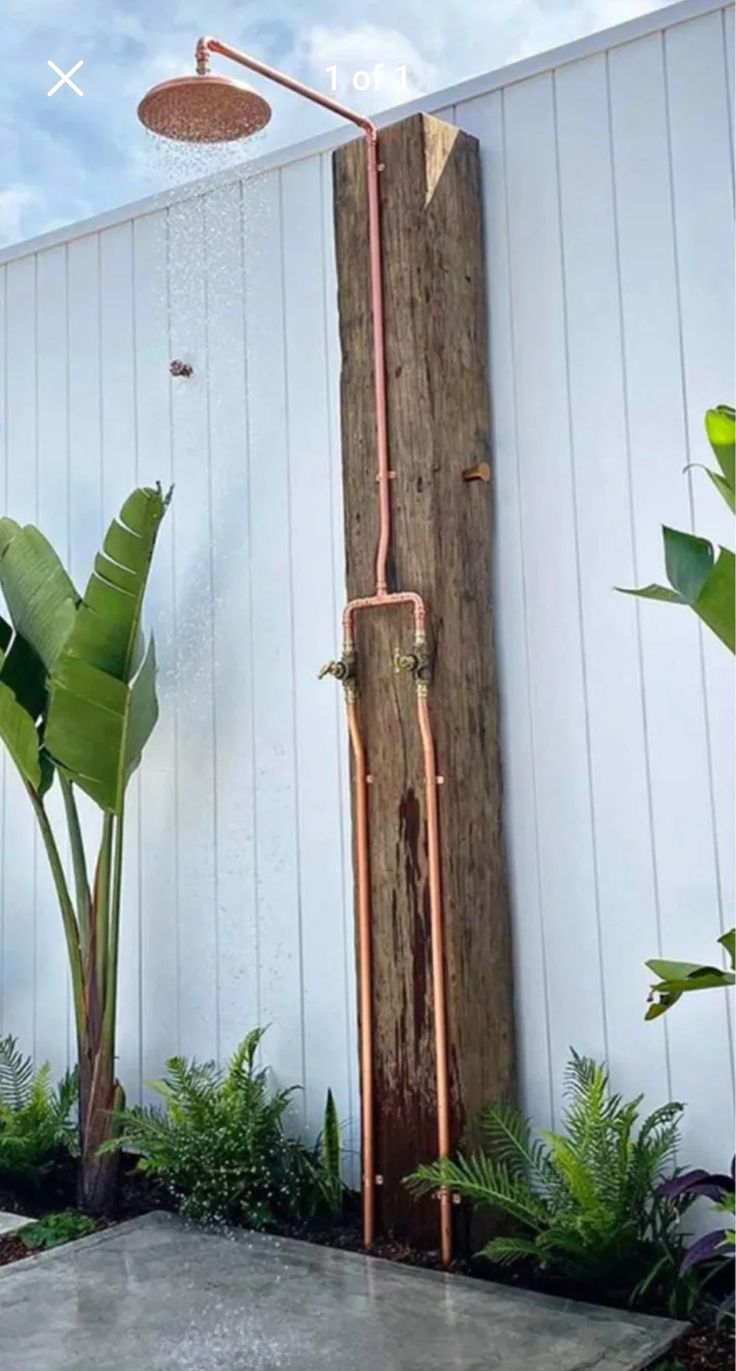 an outdoor shower in the middle of a garden with trees and plants around it, next to a white wall