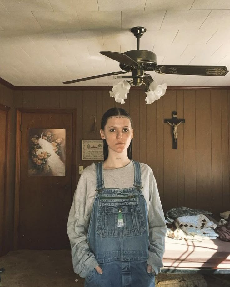 a woman wearing overalls standing in front of a bed with a cross hanging from the ceiling