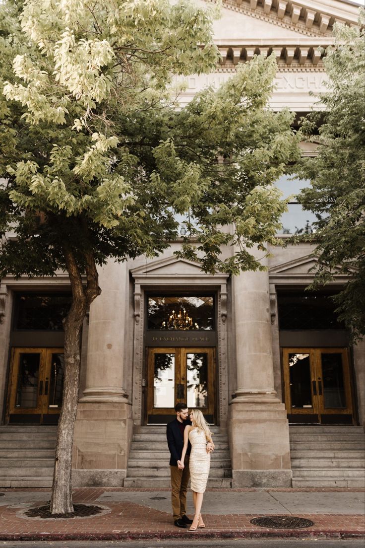 a man and woman standing in front of a large building with trees on the sidewalk