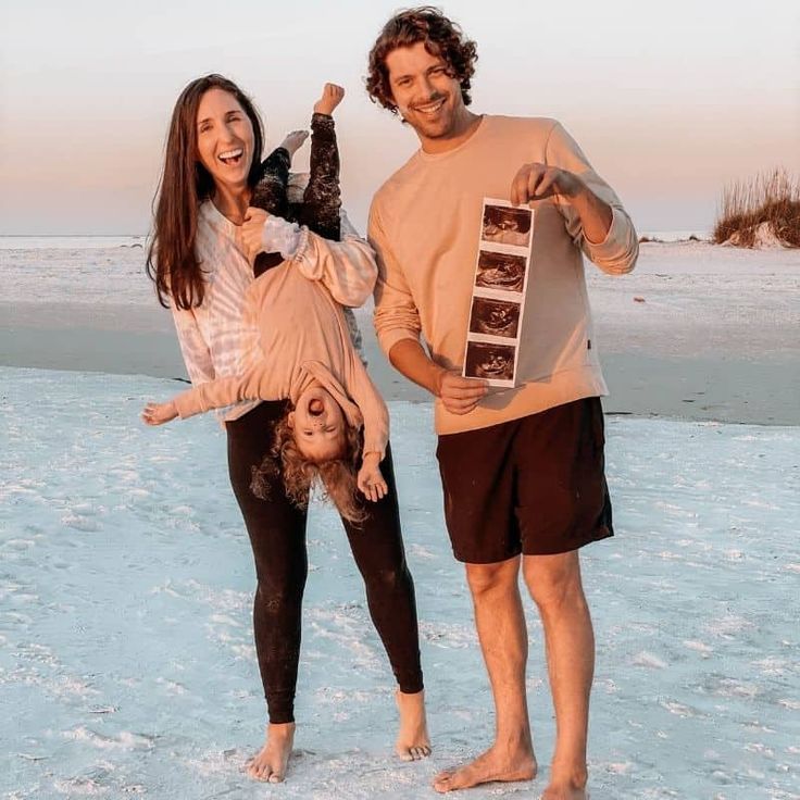 a man and woman standing on the beach with a child upside down in front of them