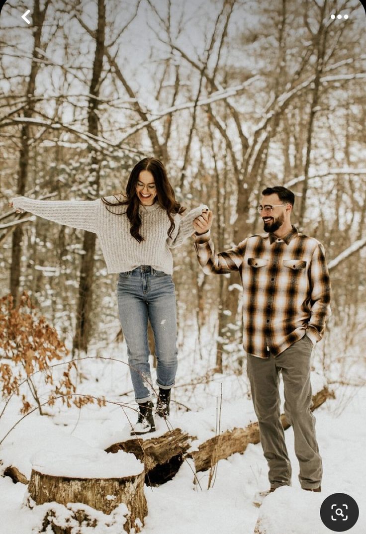 a man and woman standing on a log in the snow with their arms spread out