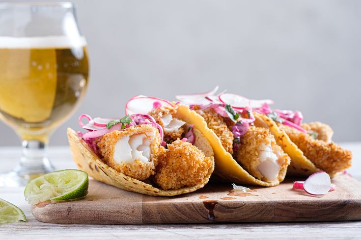 three fish tacos on a cutting board next to a glass of beer