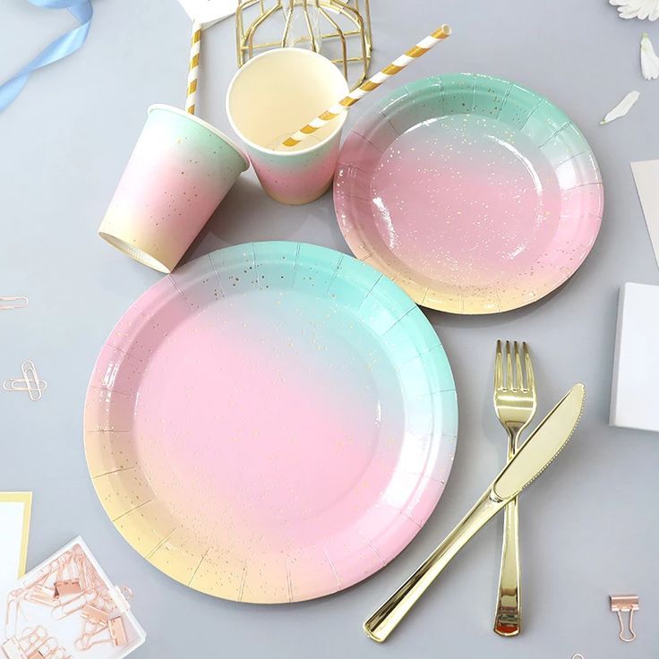 a table set with pink and blue plates, silverware and gold cutlery on it