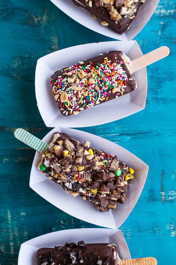 three ice cream pops with chocolate and sprinkles in paper containers on a blue table