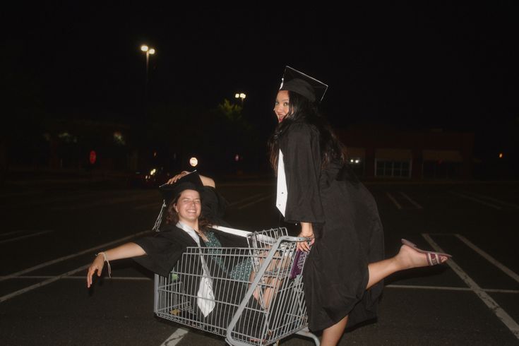 two women in graduation gowns are pushing a shopping cart with a graduate on it