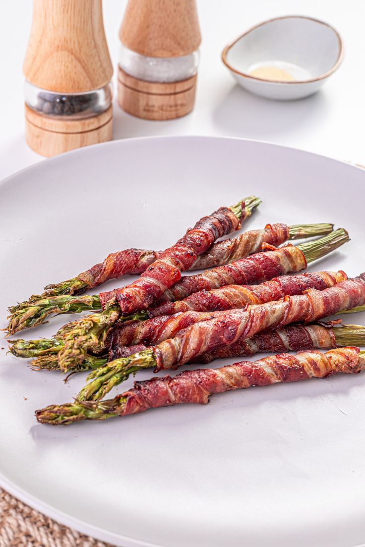 asparagus wrapped in bacon sitting on a white plate next to a salt and pepper shaker