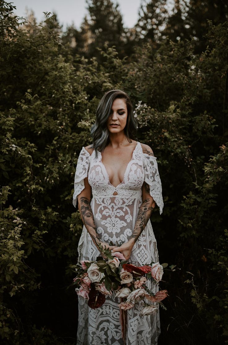 a woman standing in front of some trees wearing a white dress and holding a bouquet