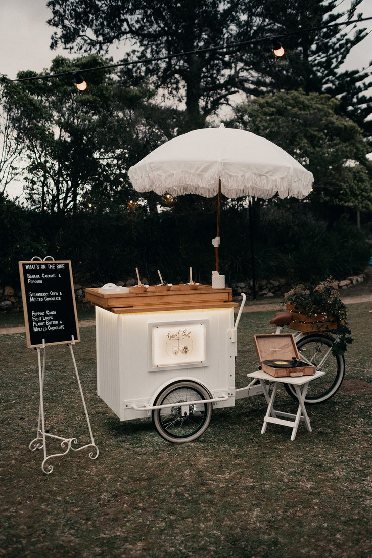 an ice cream cart is set up in the grass with an umbrella and two chairs