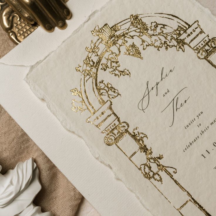 an ornate wedding card with gold foil on it next to a pair of bronze scissors