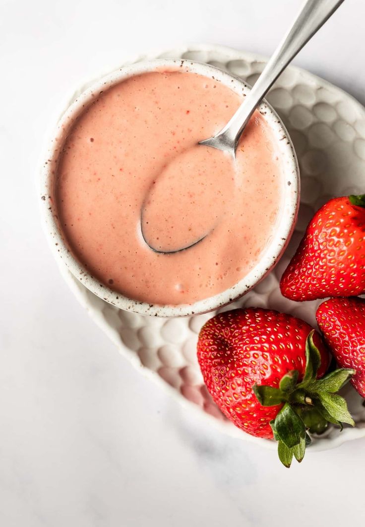 two strawberries and a bowl of strawberry smoothie on a white marble counter top