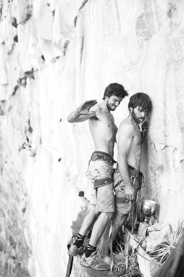 two young men climbing up the side of a mountain, one is holding onto another man's shoulder