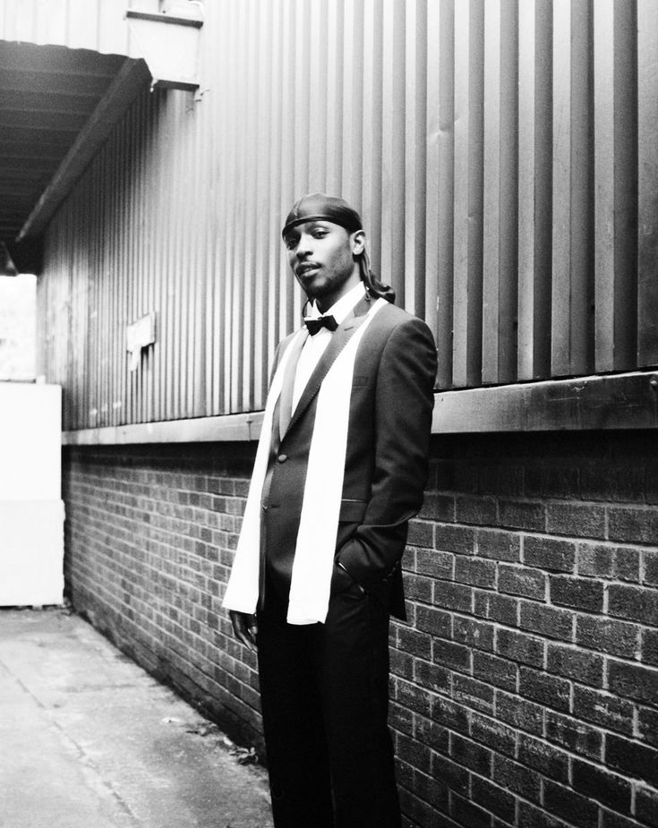 a man in a suit and bow tie leaning against a brick wall with his hands on his hips