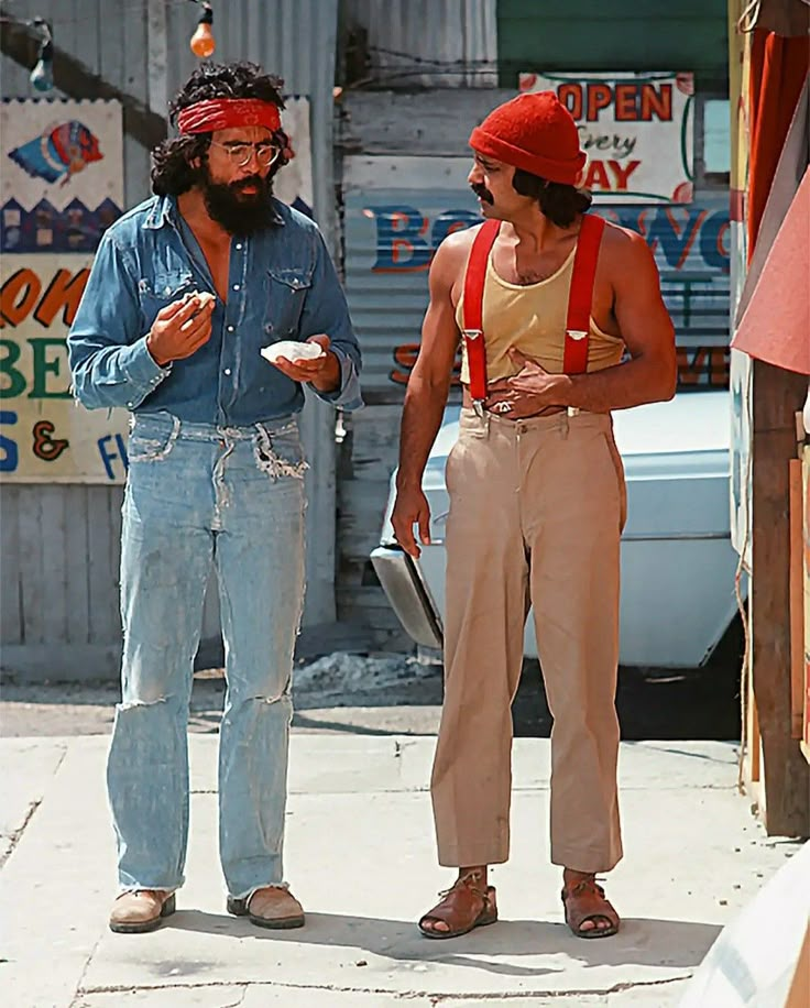 two people standing on the sidewalk eating food and one is wearing a red bandana