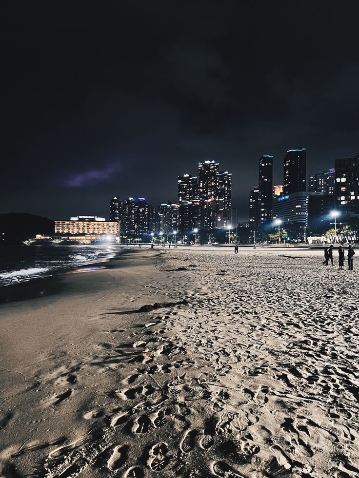 people walking on the beach at night with city lights in the backgroung