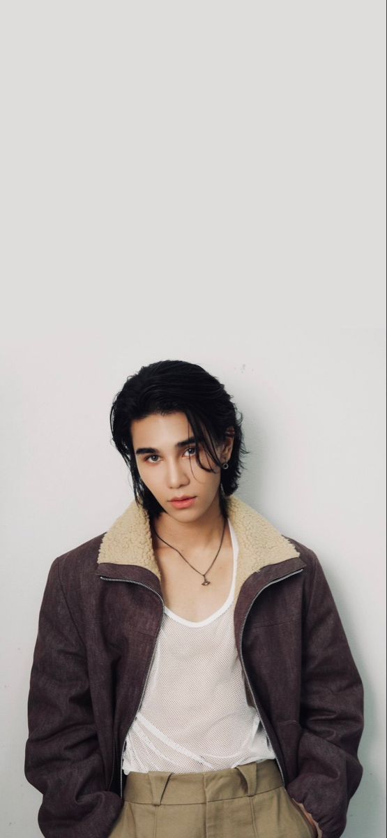 a young man wearing a brown jacket and khaki pants standing in front of a white wall