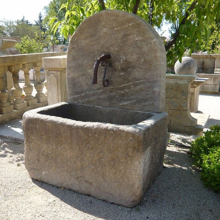 a stone fountain with a faucet in the middle surrounded by trees and bushes