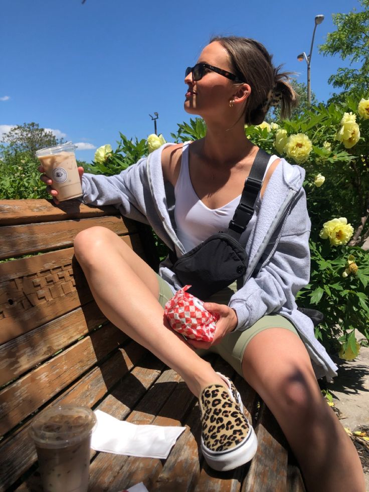 Girl sitting on bench with an ice chai tea latte in one hand and a sandwich in a wrapper in the other. Looking off to the side wearing a black lululemon Fanny pack across her chest, wearing green lululemon tight biker shorts, a white lululemon align tank top, and an oversized grey zip up sweater that is undone, with cheetah print vans slides Lululemon Fanny Pack Outfit, Black Fanny Pack Outfit, Crossbody Fanny Pack Outfit, Hip Pack Outfit, Fanny Pack Outfit Summer, Green Biker Shorts Outfit, Align Tank Outfit, Fanny Pack Outfit Street Styles, Fanny Pack Aesthetic