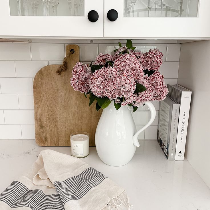 pink flowers are in a white pitcher on the kitchen counter next to a cutting board