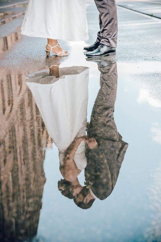 a man and woman standing next to each other holding hands in the rain with their reflection on the ground