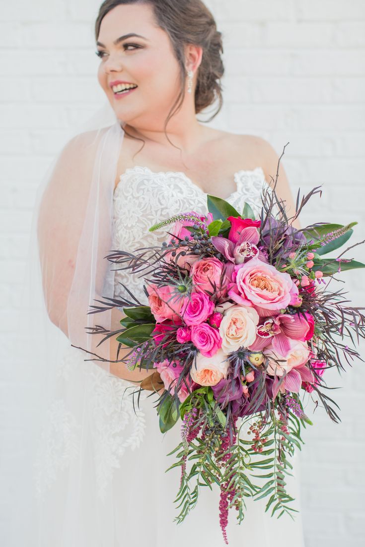 a bride holding her bouquet in front of a white brick wall with greenery and pink flowers